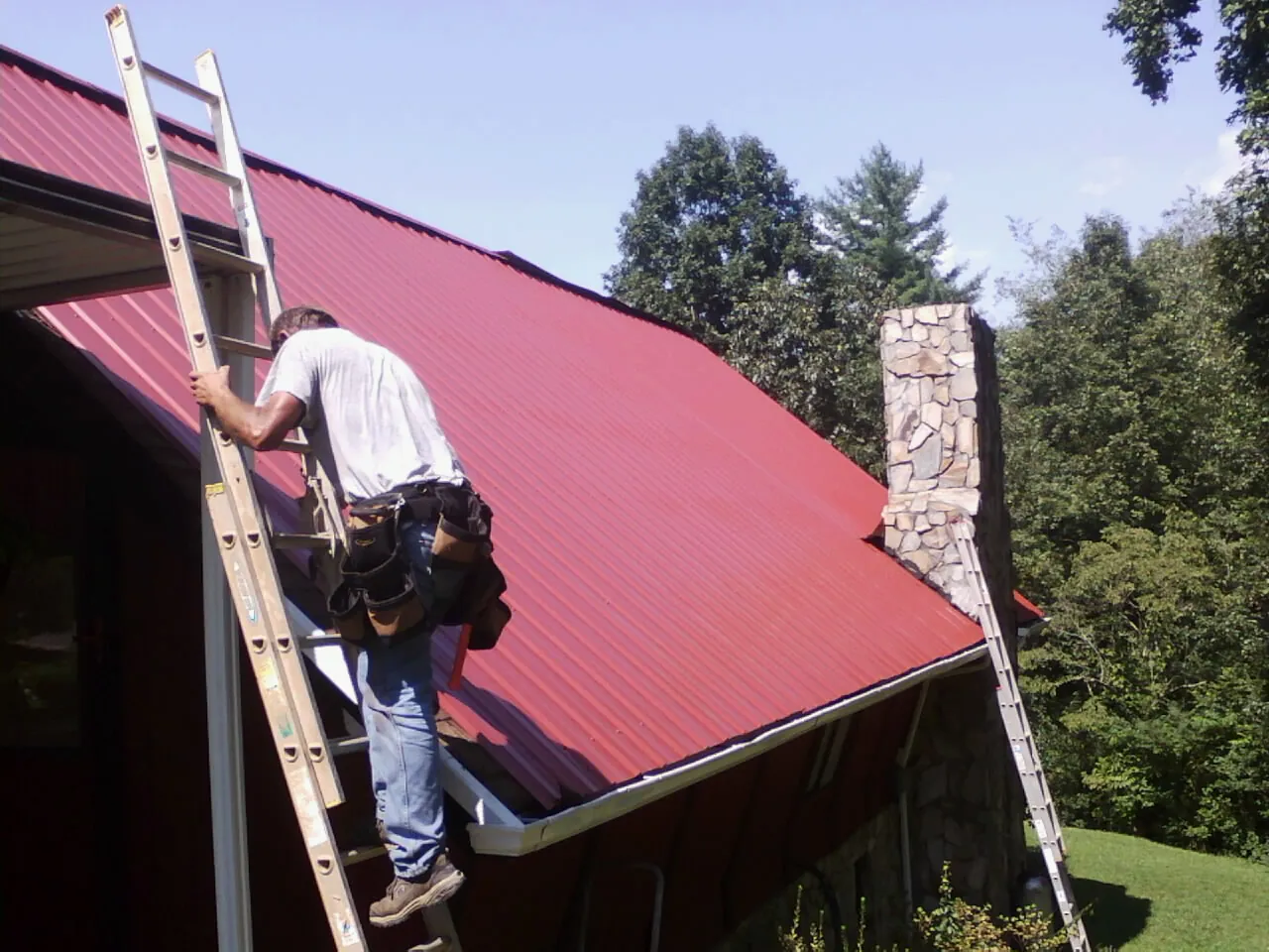 Roofer on a ladder beside a red metal roof and stone chimney — Soffit & Fascia Repair by Roofing Brothers in Grant